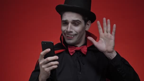 吸血鬼男子与血和尖牙之间的黑色半边之间的服装挥手和笑 同时有一个视频电话智能手机隔离红墙 — 图库视频影像