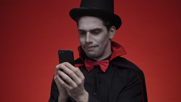 怒气冲冲的吸血鬼男人 血气方刚 身穿黑色半身长牙服 鬼鬼祟祟地笑着 一边用智能手机与红墙隔离聊天 — 图库视频影像