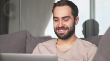 Memnun bir genç adam dizüstü bilgisayarında bir şey izliyor. Oturma odasındaki kanepede oturuyor.