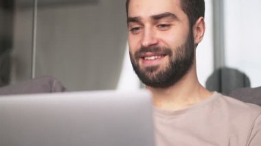 Mutlu genç bir adamın yakın plan görüntüsü dizüstü bilgisayarını evde oturma odasındaki kanepede otururken kullanıyor.