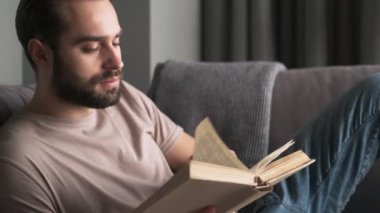 Yakışıklı bir genç adam evde, oturma odasındaki kanepede oturmuş kitap okuyor.