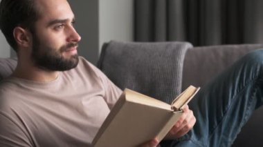 Sakin bir genç adam evde, oturma odasındaki kanepede oturmuş kitap okuyor.