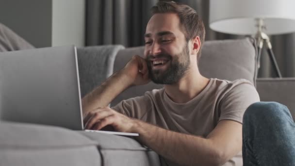 幸せな若い男がタイプしています彼のラップトップに座って自宅のリビングルームで — ストック動画