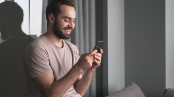 Ein Fröhlicher Junger Mann Sitzt Mit Seinem Smartphone Hause Wohnzimmer — Stockvideo