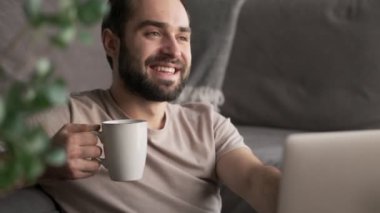 Çekici genç bir adam evinde otururken dizüstü bilgisayarını kullanarak bir fincan çay tutuyor.