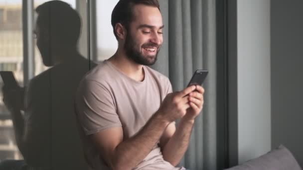 Ein Lachender Junger Mann Sitzt Mit Seinem Smartphone Hause Wohnzimmer — Stockvideo