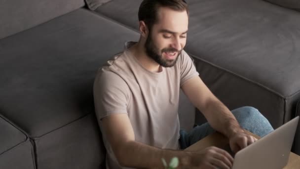 一位面带微笑的年轻人正坐在客厅里用他的笔记本电脑 — 图库视频影像
