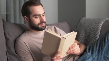 Çekici bir genç adam evde, oturma odasındaki kanepede oturmuş kitap okuyor.