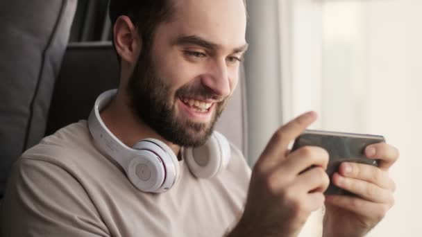 幸せな感情的な若い男が自宅のリビングルームに座っている間 彼のスマートフォンでゲームをしています — ストック動画