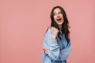 Uzun kahverengi saçlı, gülümseyen ve pembe arka planda izole edilmiş boş bir alanı işaret eden çekici bir genç kadın.