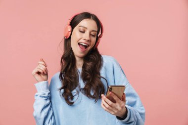 Uzun kıvırcık esmer saçlı pembe arka planda dikilip kulaklık ve cep telefonuyla müzik dinleyen uzun ve neşeli bir kızın portresi.