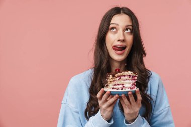 Pembe arka planda izole edilmiş pastayı tutarken dudaklarından hoşlanan genç bir kadının görüntüsü