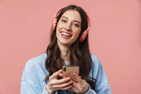 在粉色背景下使用耳机和手机时高兴地大笑的年轻女性的照片 — 图库照片
