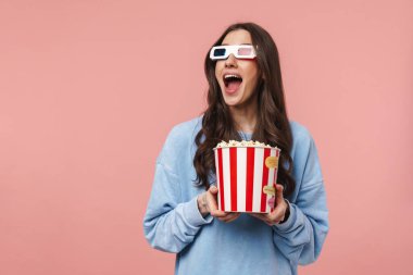 Uzun kıvırcık esmer saçlı, pembe arka planda yalnız duran, 3D gözlük takan, elinde patlamış mısır kovası tutan güzel bir kızın portresi.