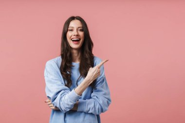 Uzun kahverengi saçlı, gülümseyen ve pembe arka planda izole edilmiş boş bir alanı işaret eden çekici bir genç kadın.