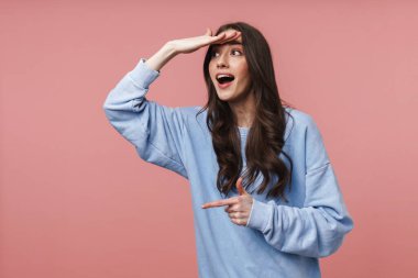 Uzun kahverengi saçlı çekici bir genç kadın görüntüsü kenara bakıyor ve pembe arka planda izole edilmiş boş bir alanı işaret ediyor.