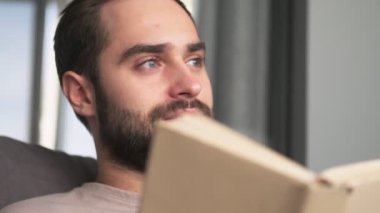 Çekici bir gencin yakın plan görüntüsü oturma odasındaki kanepede oturmuş kitap okuyor.