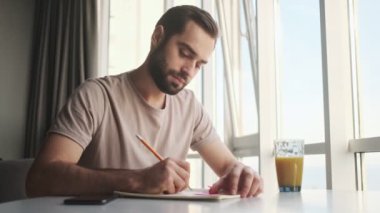Odaklanmış genç bir adam evdeki masada oturmuş bir şeyler yazıyor.