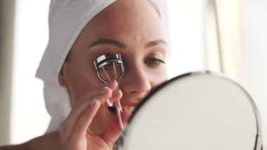 Evdeki neşeli genç kadın kirpiklerini kıvıran aynaya bakarak makyaj yapıyor.