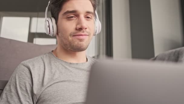 ヘッドフォンでビデオ通話で話すラップトップコンピュータを使用して自宅で屋内で男を喜ばせます — ストック動画