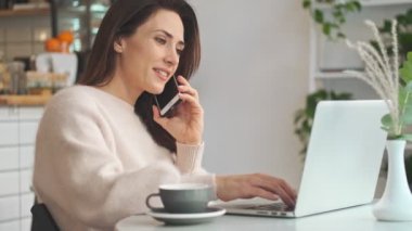 Hoş bir genç bayanın yan görüntüsü telefonda konuşuyor ve kafede otururken dizüstü bilgisayarında daktilo yazıyor.