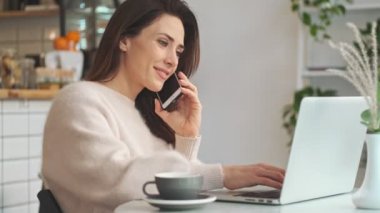Gülüp geçen genç bir kadının yan görüntüsü telefonla konuşuyor ve kafede otururken dizüstü bilgisayarında daktilo yazıyor.