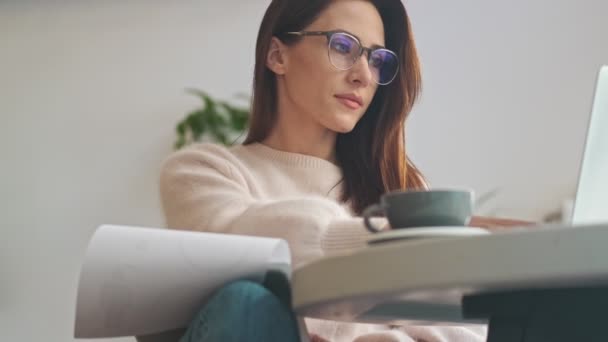 一位漂亮而沉着的年轻女士带着眼镜 一边在室内的咖啡店里与笔记本电脑一起工作一边做笔记 — 图库视频影像