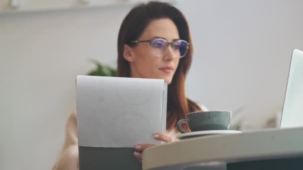 一个戴眼镜的漂亮的年轻女人在屋里看手提电脑时正在做笔记 — 图库视频影像