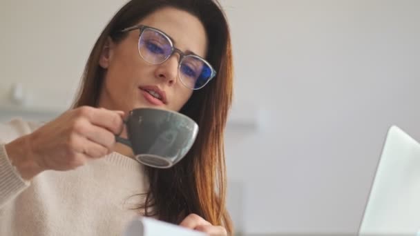 白い居心地の良いカフェで働いている間 眼鏡をかけた素敵な若い女性がコーヒーを飲んでいます — ストック動画