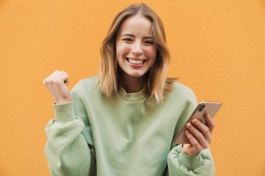 Mutlu sarışın kadın portresi cep telefonu kullanıyor ve sarı arka planda izole edilmiş bir jest yapıyor.