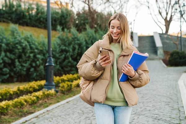 公園を歩きながら運動本を持って携帯電話を使っている笑顔の若い女性の肖像画 — ストック写真