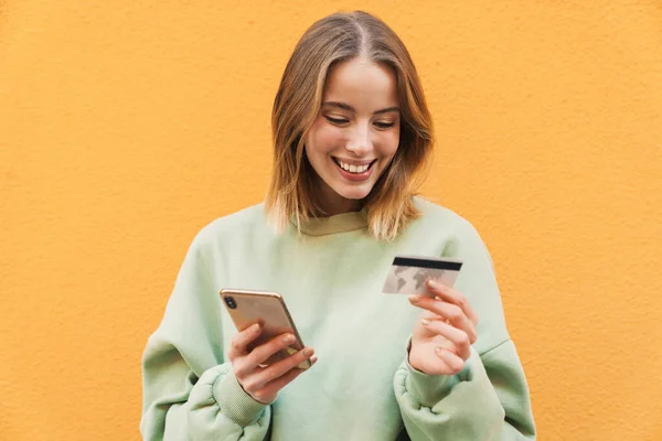 笑顔ブロンド女性の肖像画保持クレジットカードと使用携帯電話絶縁上の黄色の背景 — ストック写真