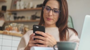 Gözlüklü güzel bir kadın beyaz kafeteryada akıllı telefonunu kullanıyor.
