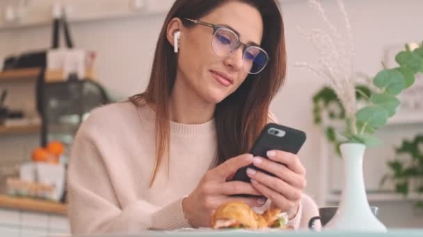 心地よいカフェに座っている朝食時に イヤホン付きの女性がスマートフォンを使用しています — ストック動画
