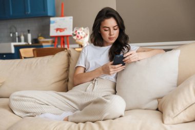 Güzel, genç, esmer bir kadın evdeki kanepede dinleniyor. Cep telefonu kullanıyor.