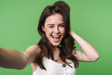 Heyecanlı çekici bir kadının yeşil arka planda izole edilmiş, gülümseyen ve selfie çeken görüntüsü.