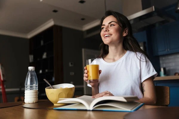 迷人的 面带微笑的年轻黑发女人一边看书 一边吃着健康的早餐 一边坐在厨房桌子旁 手里拿着一杯橙汁 — 图库照片