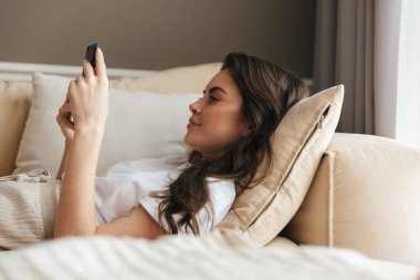 Güzel gülümseyen genç esmer kadın evdeki koltukta dinleniyor, cep telefonu kullanıyor.