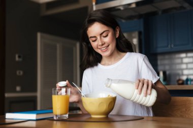 Çekici gülümseyen genç esmer kadın mutfak masasında oturup mısır gevreğine süt dökerken sağlıklı bir kahvaltı yapıyor.