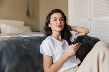 Çekici, gülümseyen, esmer bir kadın. Yerde otururken cep telefonu kullanıyor. Yatak odasında yatağa yaslanıyor.