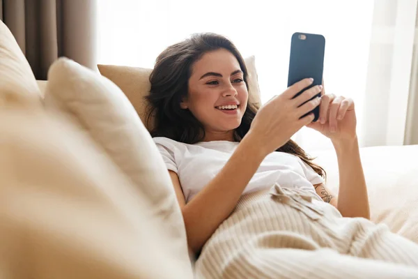 Красивая Улыбающаяся Юная Брюнетка Расслабляется Дома Диване Используя Мобильный Телефон — стоковое фото
