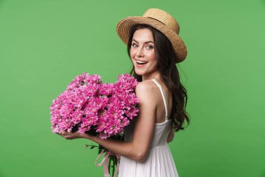 Hasır şapkalı neşeli güzel bir kadın resmi. Gülümsüyor ve yeşil arka planda saklanmış çiçekleri tutuyor.