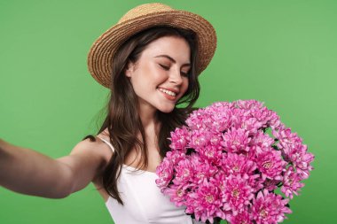 Yeşil arka planda izole edilmiş selfie çekerken elinde çiçekle saman şapkalı neşeli bir kadın görüntüsü