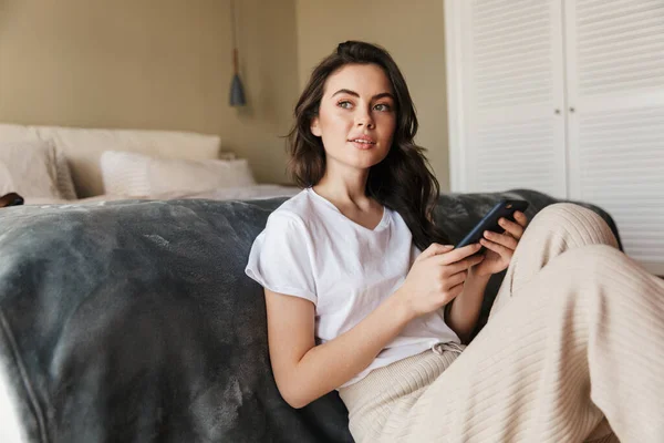 Vonzó Mosolygós Fiatal Barna Használja Mobiltelefon Miközben Padlón Támaszkodva Ágyra — Stock Fotó