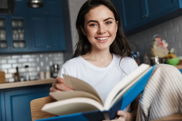 Atraente Sorrindo Jovem Morena Lendo Livro Enquanto Sentado Uma Cozinha — Fotografia de Stock