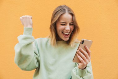 Mutlu sarışın kadın portresi cep telefonu kullanıyor ve sarı arka planda izole edilmiş bir jest yapıyor.