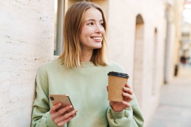 Gülümseyen genç bir kadının portresi. Kahve içerken ve sokakta yürürken cep telefonu kullanırken.