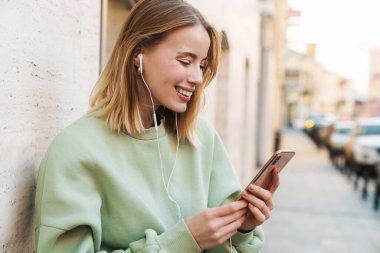 Şehirde yürürken kulaklık ve cep telefonu kullanan gülümseyen genç bir kadının portresi