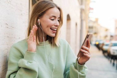 Şehirde el sallarken kulaklık ve cep telefonu kullanan gülümseyen genç bir kadının portresi.