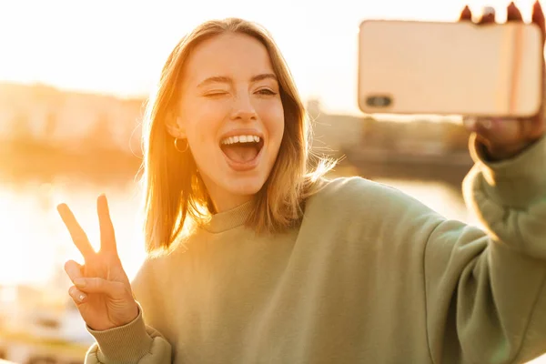 Portrait of cheerful woman gesturing peace sign and winking while taking selfie on cellphone at promenade